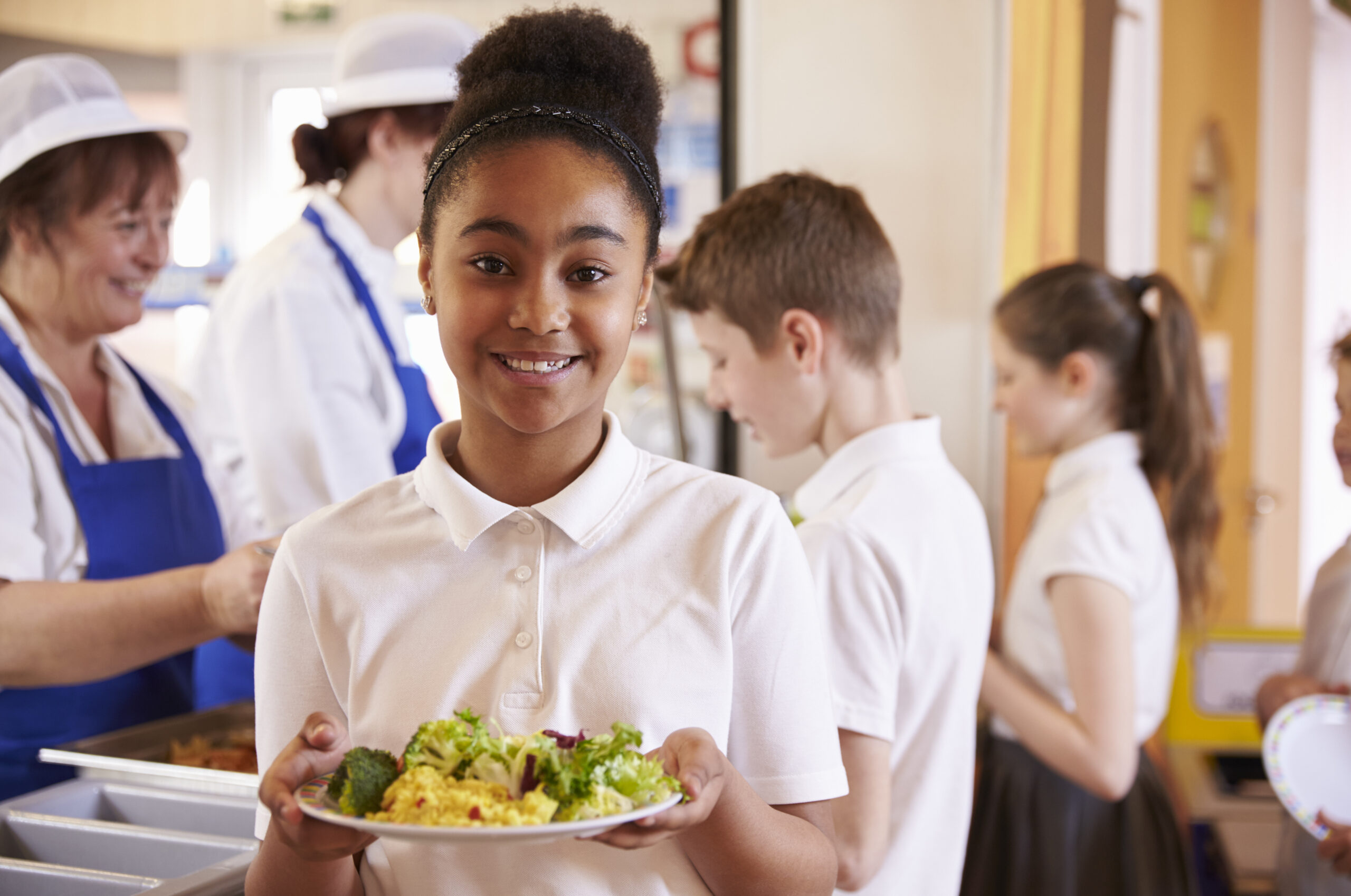 a foto mostra uma menina negra segurando um prato com comida. atrás delas outras duas crianças estão na frente de uma mesa com comidas sendo servidas por duas mulheres que usam avental e touca