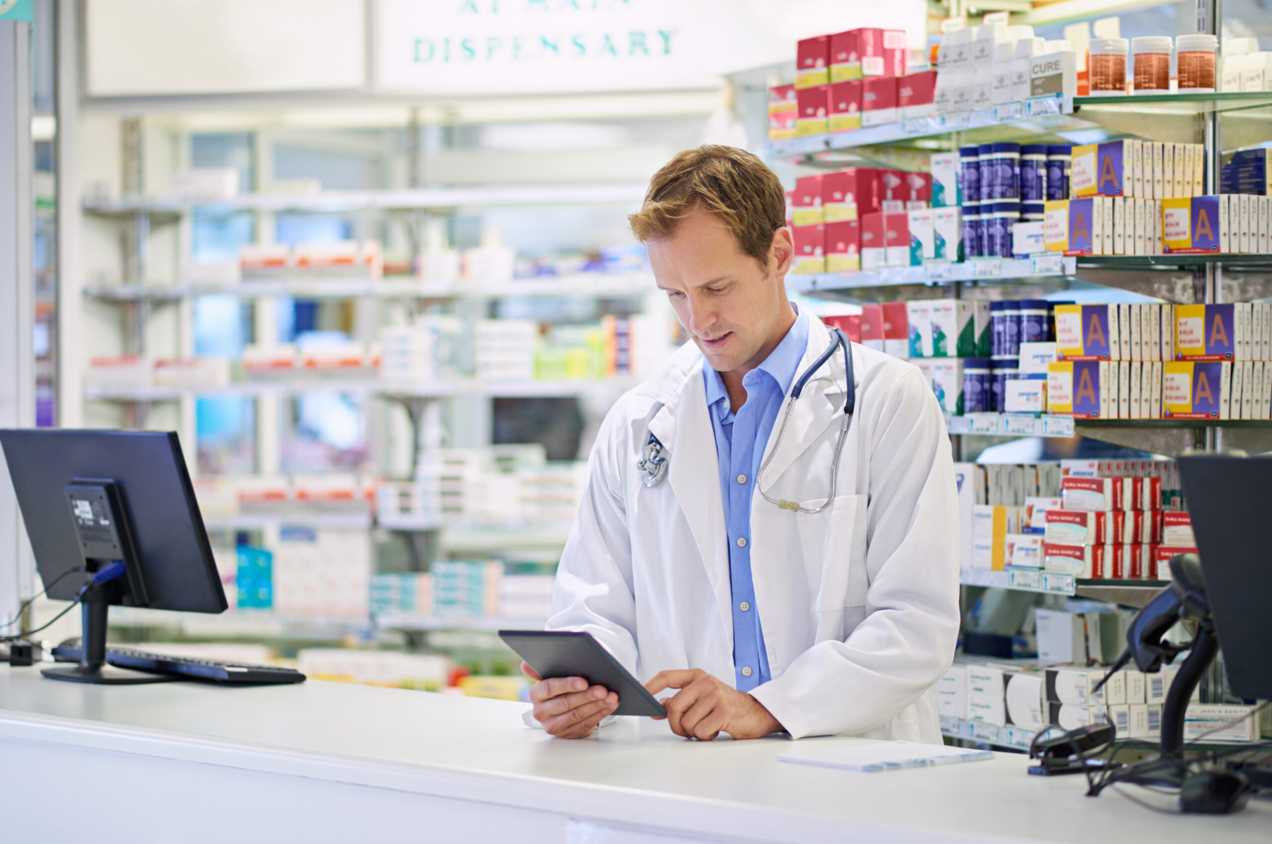a foto mostra uma farmacêutico olhando um tablet atrás de um balcão. ao fundo, temos prateleiras de medicamentos