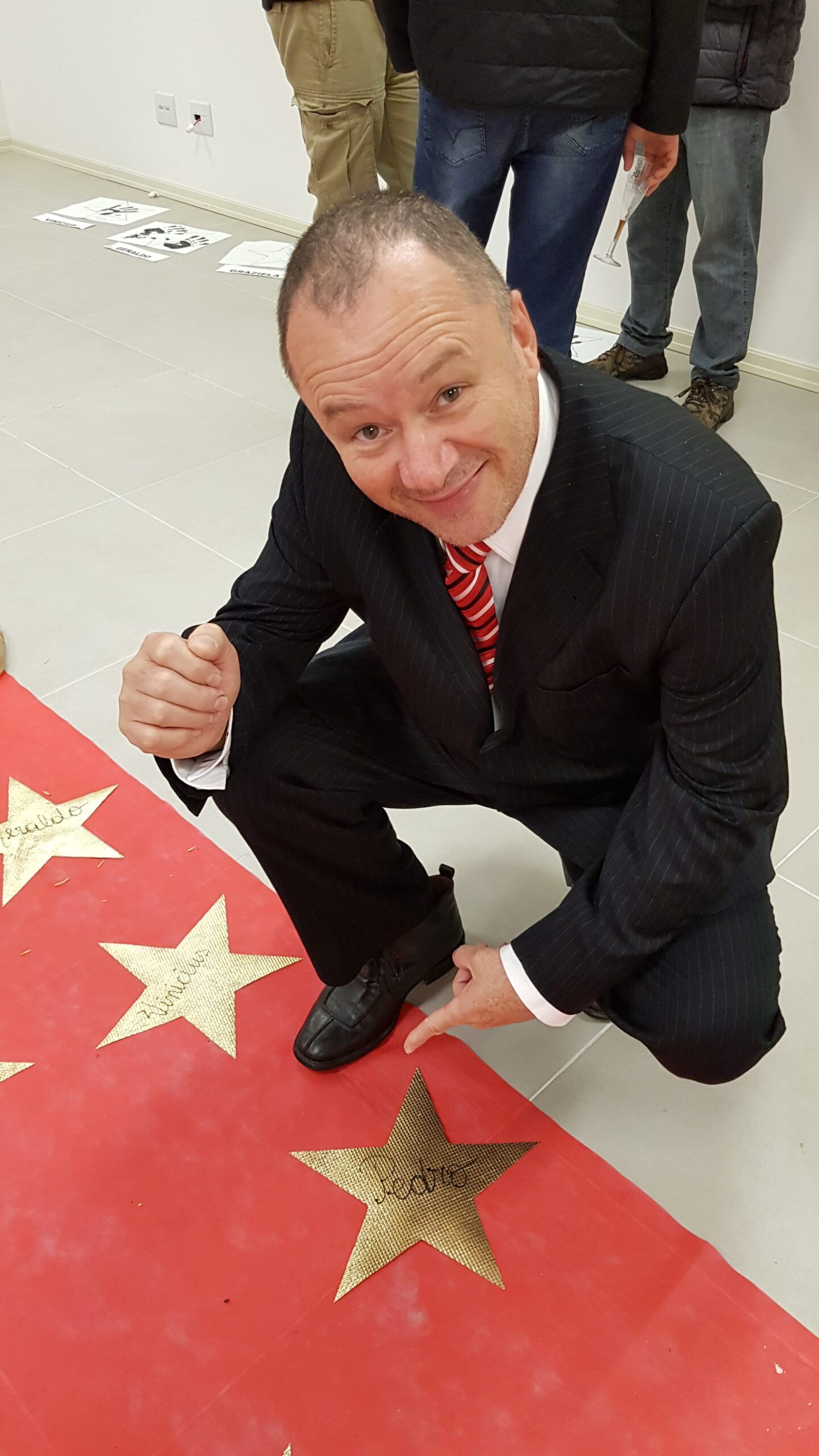 a foto mostra um homem, de terno, agachado, apontado para o chão que tem um corredor vermelho com estrelas douradas com nomes de pessoas