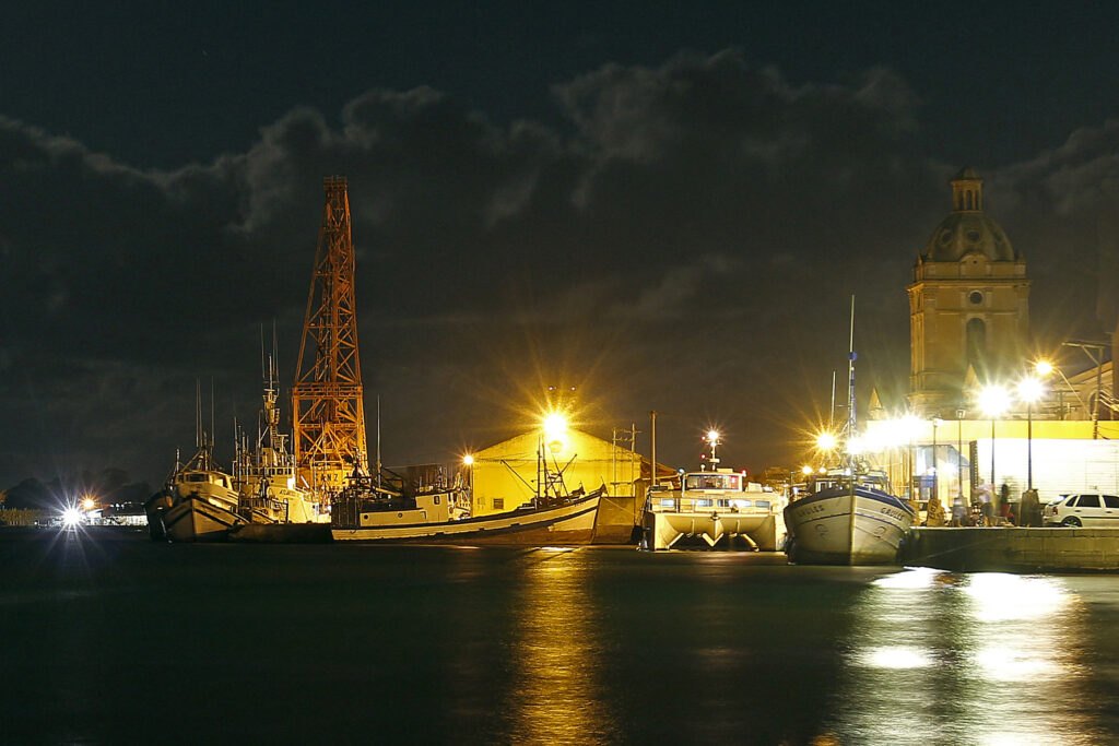 Foto do Porto de Rio Grande à noite, com o cais iluminado, barcos atracados, e uma torre de igreja ao fundo.