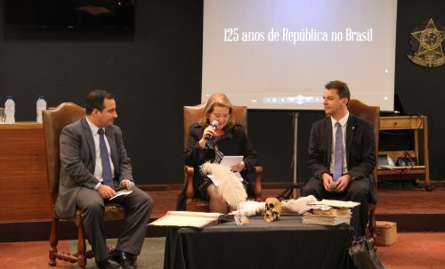 Três debatedores sentados. No meio a desembargadora federal Marga Inge Barth Tessler entre duas outras autoridades: Dr.Eduardo Tonetto Picarelli à esquerda e Dr. Antônio César Bochenek à sua direita, os quais conduziram solenidade. Atrás uma projeção com o título 125 anos de República no Brasil.