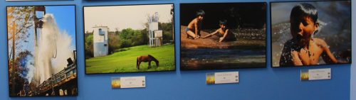 Parede com 4 quadros uma ao lado do outro horizontalmente: O primeiro tem a imagem de um maquinário grande em céu aberto; dele , sai um jato de água. Mais à esquerda da foto há uma parte de uma árvore. Um homem bem embaixo observa o jato de água. No segundo quadro temos a foto/imagem de um cavalo pastando em um gramado bem verde; ao fundo há arbustos e duas construções arredondadas, uma de cada lado da foto. Terceiro quadro há duas crianças à beira de um rio: uma em uma pedra , a outra no lado desta mas já se enconstando na água. Quarto quadro há uma criança se banhando em um rio, foto do tronco pra cima.