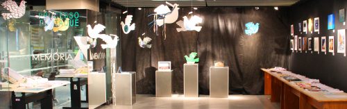 Foto de uma parte do espaço do memorial . Há pássaros de papel pendurados nos forros do teto. Ao fundo da foto, a parede está forrada de tecido preto, e há três colunas pequenas com objetos expostos em cima de cada. No lado direito da foto , uma mesa grande com figuras/documentos sobre ela, bem como na parde preta rente a esta mesa. No lado esquerdo da foto (lado onde fica a entrada do memorial ) há uma mesa e livros/documentos sobre ela.