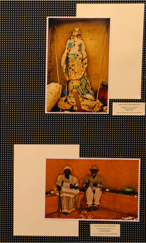 Duas fotos na imagem : Figura 1: foto da imagem de uma santa (parece ser Iemanjá) aparentemente em local aberto , como se ela estivesse numa grutinha para visita de adeptos; a pintura está bem descascada (era azul) e a imagem bem danificada. Há uma pequena concha rente ao seu vestido. Figura 2: Casal de bonecos negros sentados, vestidos conforme a cultura e roupa de sua época: A mulher de vestido longo com rendas, um turbante na cabeça, guia no pescoço, cajado numa mão e um objeto na outra; o homem, com camisa cinza, calça branca , sandalhas, chapéu, guia no pescoço; também com um cajado numa mão e objeto na outra. Estão sentados numa mureta rente a uma parede (que serve como um "sofá"); ao lado de cada um há uma xícara branca.
