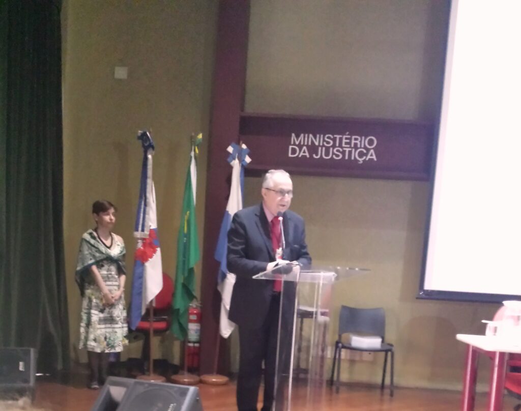 Sr. Jaime Antunes da Silva, então Diretor do Arquivo Nacional, em pé falando em um microfone (o público para o qual ele se dirige não aparece na foto). Atrás dele há uma mulher em pé ao lado de três bandeiras não hasteadas e escrito "Ministério da Justiça".