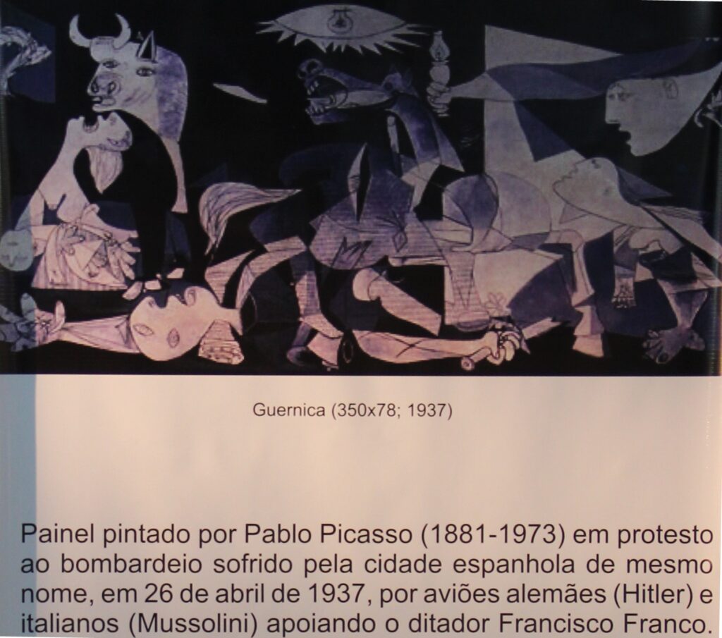 Foto do painel de Picasso com descrição embaixo. Título: Guernica. Descrição: "Painel pintado por Pablo Picasso (1881-1973) em protesto ao bombardeio sofrido pela cidade espanhola de mesmo nome, em 26 de abril de 1937, por aviões alemães (Hitler) e italianos (Mussolini) apoiando o ditador Francisco Franco.