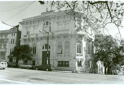 foto semi-diagonal do "Palacinho" , prédio histórico localizado na avenida Cristovão Colombo (numa esquina) . Foto antiga, em preto e branco. Prédio : cor clara; há carros estacionados na frente; ao lado do prédio, uma rua com árvores e carros estacionados.