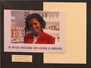 Título: “A etnia incluída em cores e odores”. Foto de uma mulher (do tronco para cima) afrodescendente em local onde há frascos cujo conteúdo é um líquido colorido. Estes frascos estão em estantes na parede ao lado e atrás dela. A mulher está sorrindo e veste um blazer vermelho.