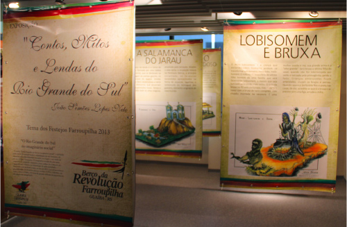 Banners pendurados ao longo do espaço do memorial; primeiro banner está escrito "Contos, Mitos e Lendas do Rio Grande do Sul"; no segundo: "Lobisomem e Bruxa" e um texto que segue abaixo (não possível de ler) seguido de uma imagem; mais atrás, ao fundo da foto, “A Salamanca do Jarau” e um texto que segue abaixo (não possível de ler também) seguido de uma imagem.