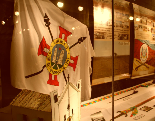 Imagem de uma parte da vitrine exposta: O fundo da vitrine é a parede. conteúdo da vitrine que aparece na foto: à esquerda da imagem , a bandeira da cidade de Viamão / RS. Abaixo da bandeira , há uma maquete de uma igreja branca , com uma cruz em seu topo ; ao lado , lanças expostas na horizontal sobre uma mesa / apoio forrado com pano branco. Na parede há cartazes /banners com símbolos gaúchos e textos.