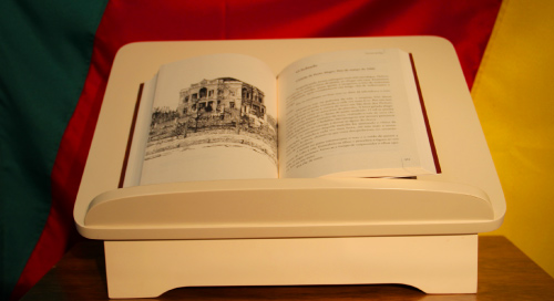 Um livro aberto sobre um apoio branco. Apoio que deixa o livro inclinado para melhor observar /ler. Página esquerda, uma foto em preto e branco de uma construção ; página à direita , um texto - não legível na foto.