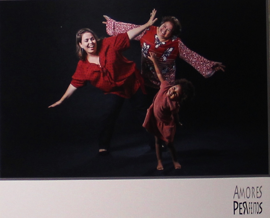 Casal homoafetivo feminino, uma à frente da outra, fazendo movimento de "aviãozinho" junto com sua filha pequena ( 3 anos de idade), mais à frente. Todas estão sorrindo.