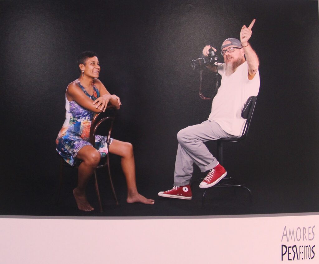 Casal sentado em cadeiras ; à esquerda uma mulher afro-brasileira e à direita um homem branco com barba, segurando uma câmera fotográfica com a mãe esquerda, ambos interagindo alegremente um com outro.