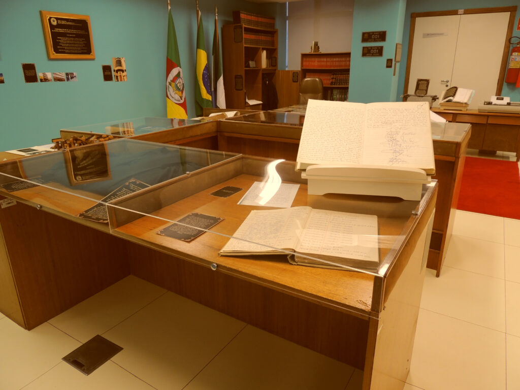 Espaço do memorial Rio Grande: mesas com expositores de vidro embutidos no topo. Há 3 mesas em forma de U; dentro desses expositores, processos, documentos e descrições a respeito. Na parede à esquerda, de cor azulada, há pequenas fotos e uma placa pendurada (não é possível ler o que está escrito). Logo após, três bandeiras em mastros: a do Rio Grande do Sul, a do Brasil e a de Rio Grande. Ainda junto à parede, uma estante com livros. Na parede que segue, oposta à foto, uma estante menor, com livros e uma porta dupla de entrada; ao lado desta um extintor de incêndio. Há uma outra mesa próxima à entrada de frente com um processo exposto; ao lado deste uma máquina de escrever. Observa-se um tapete vermelho no piso claro.