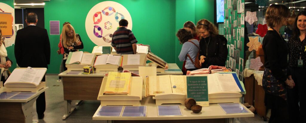 Espaço do memorial com processos expostos em mesas diversas e pessoas circulando ao redor. Fundo da foto : uma parede verde com um símbolo redondo e desenhos coloridos dentro.