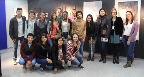 Grupo de jovens adolescentes junto com uma servidora, pousando para foto. 12 estão em pé, um ao lado do outro; 4 estão abaixados/acocados à frente - mais para o lado esquerdo. Atrás, parede preta com figuras /artes referentes à exposição.