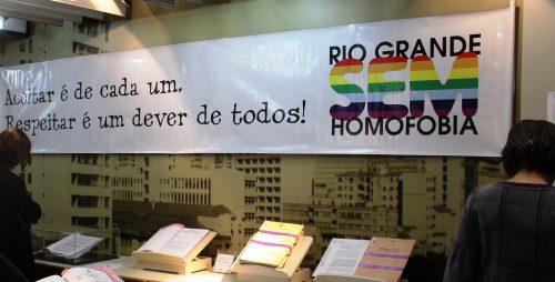 Cartaz horizontalmente comprido escrito : "Aceitar é de cada um. Respeitar é um dever de todos! RIO GRANDE SEM HOMOFOBIA" . A palavra SEM está colorida (vermelho, laranja, amarelo, verde, azul, roxo,..)- Este cartaz está numa parede, e nesta há imagem de prédios. À frente, uma mesa com três processos expostos em apoios , e um em um acrílico. Nos extremos da foto (canto esquerdo e direito). Duas mulheres de costas, observando os processos. A foto capturou apenas a parte da cintura/quadril pra cima dessa mulheres. Ambas com roupas escuras.