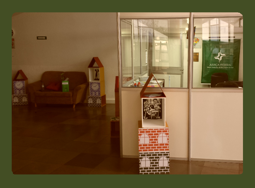 Uma sala com um gabinete dentro (com divisórias com vidros ). Dentro do gabinete se observa mesa, cadeiras e um banner da justiça federal. No espaço da sala, ao fundo da parede, há um sofá marrom com objetos em cima. Em cada lado do sofá há montagens com cubos estilizados, imitando tijolos , janelas, formando uma maquete de pequenas casas. Mais à frente, na frente do gabinete , a mesma maquete com cubos. Observa-se a figura de uma família dentro de um dos cubos que é vazado.