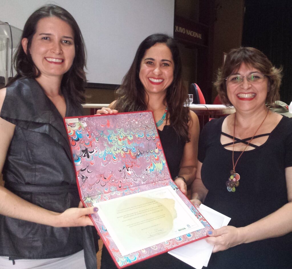 Três servidoras da Justiça Federal recebendo o Certificado Memória do Mundo em evento realizado. As três seguram uma pasta aberta , capa colorida, com o certificado impresso em um dos lados da pasta.