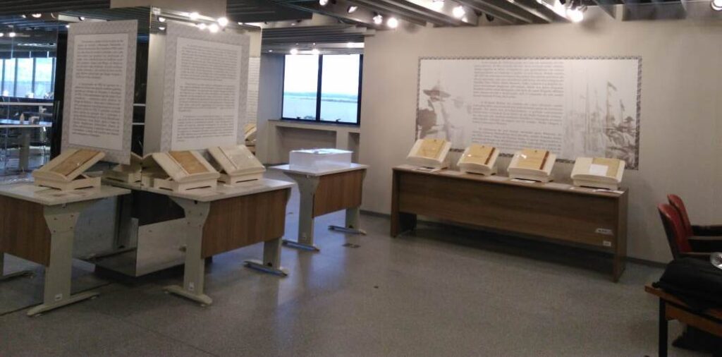 Espaço do memorial que ilustra a exposição . Há processos sobre mesas , abertos em apoiadores. Ao lado esquerdo da foto, uma coluna com adesivos e textos. No outro lado do espaço, à direita dessa coluna , mais ao fundo , há uma janela mostrando o rio Guaíba. Após, na parede do lado direito há um adesivo grande, com texto e foto.