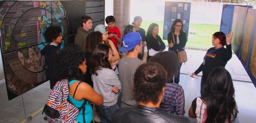 Grupo de estudantes/adolescentes (estão em 13) e duas mulheres, olhando para uma servidora e prestando atenção a sua explicação acerca da exposição. Estão em volta da servidora em forma de U; alguns estão de costas para a foto. A servidora está à direita da foto. Todos estão entre foans e cartazes com o conteúdo da exposição ( conteúdo não possível de se ler na foto.)