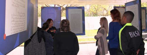 Pequeno grupo de servidores (estão em torno de 7) olhando para a servidora que está explicando a temática da exposição. Estão em volta da servidora, de costas para foto. A servidora está à esquerda da foto, mais ao fundo. Todos estão entre foans e cartazes com o conteúdo da exposição ( conteúdo não possível de se ler na foto.)