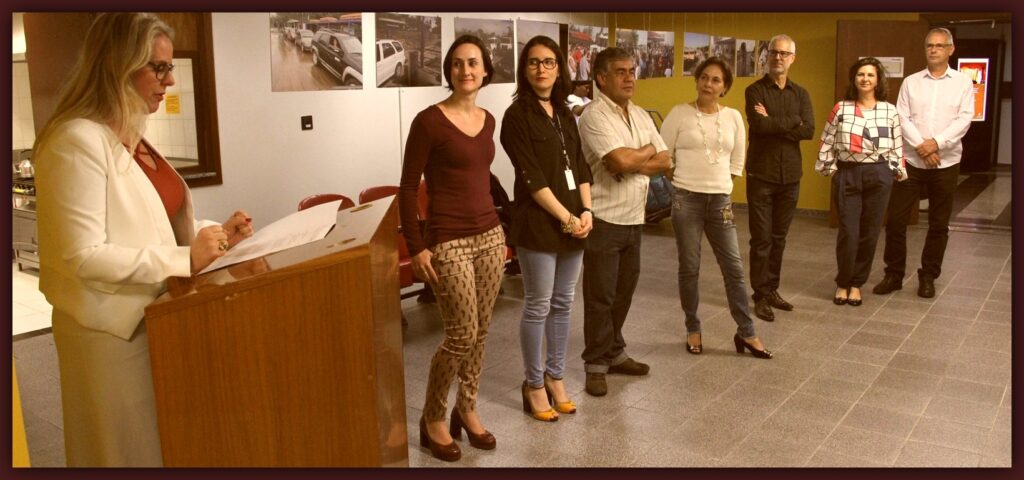 Foto com a Dra. Daniela Tocchetto Cavalheiro, à esquerda discursando , e servidores ao lado observando. Atrás destes, fotos acerca do tema da exposição, em foans pendurados .