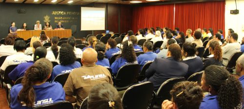 Auditório da Justiça Federal cheio, com pessoas sentadas assistindo a uma palestra; ao fundo do auditório uma bancada com uma mesa com 5 pessoas conduzindo / participando do evento.