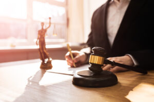 a foto mostra o tronco de um homem sentado atrás de uma mesa, vestindo terno, as mãos assinando um documento. na mesa, há um malhete e a estátua da justiça