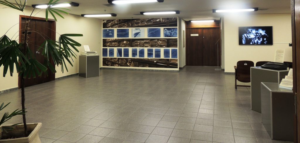 Espaço da JFRS onde ocorreu a exposição. nas paredes ao fundo do espaço , no lado esquerdo tem uma adesivo que coupa toda a parede, uma montagem com imagens do processo alternando com negativo de fotos de processo. No lado direito, uma tela passando um documentário/filme a respeito. Estão espalhados ao longo do perímetro do espaço caixas cinzas com máquinas de escrever em cima e documentos . Mais a frente , próximo de quem tirou a foto, no lado esquerdo, há uma planta.