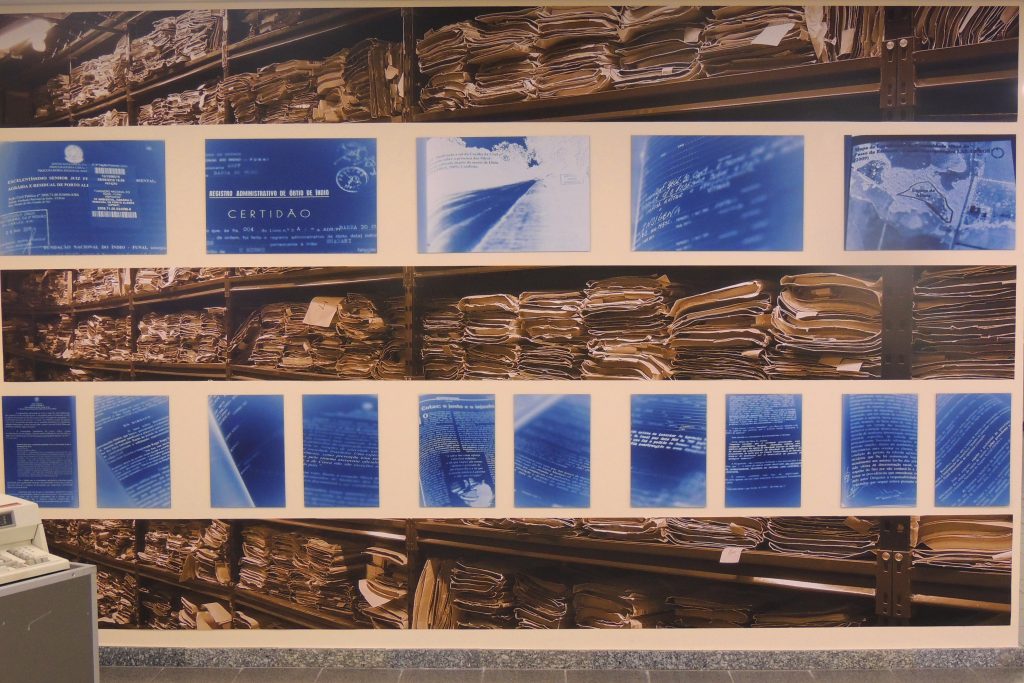 Imagens que colocaram na parede da exposição divididas em 5 linhas horizontais intercaladas ao longo de toda a parede. Na linha superior, na do meio e a última, há fotos de muitos processos em uma estante ; nas outras linhas horizontais, fotos de partes do processo e outros documentos num tom azulado.