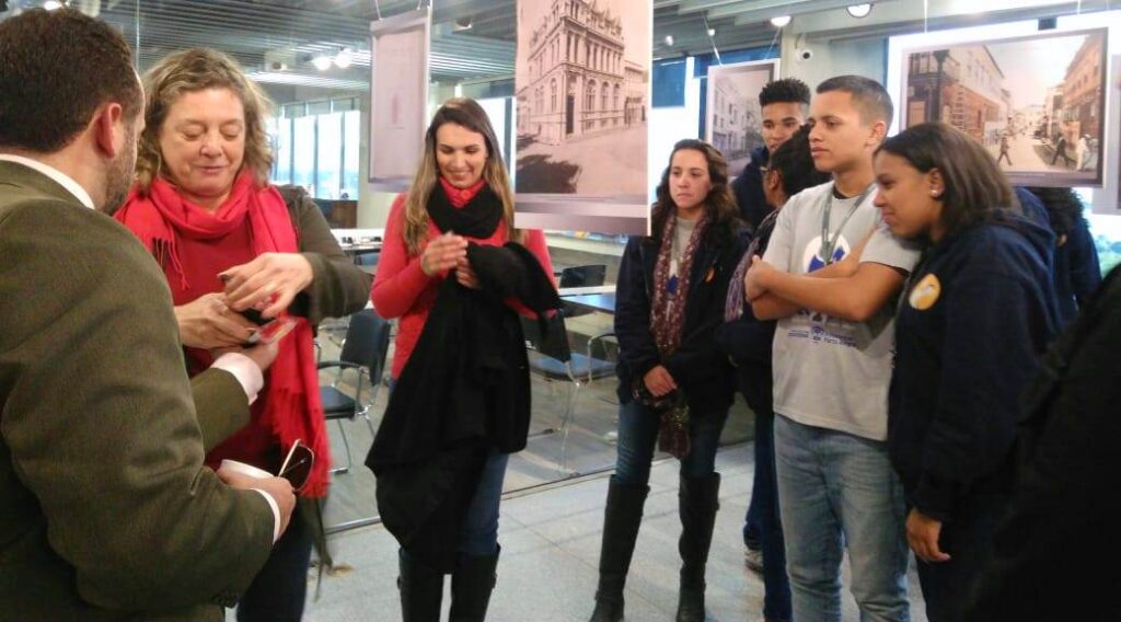 Alunos do Projeto Pescar no Espaço do Memorial junto com a servidora Adriana Bednarz. Em volta deles Foans com imagens da exposição.