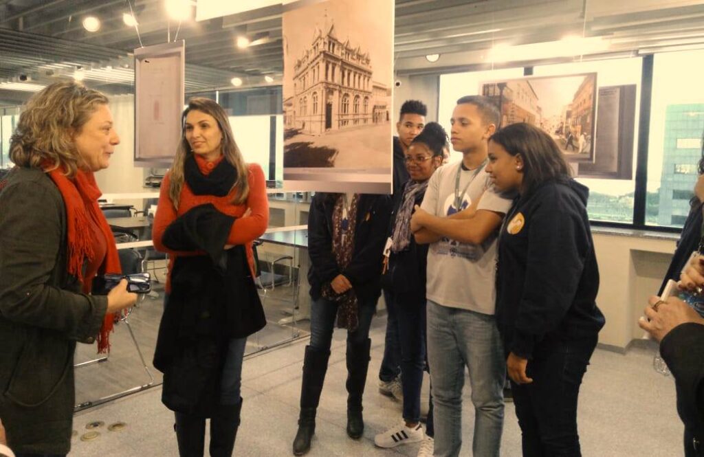 Alunos do Projeto Pescar no Espaço do Memorial junto com a servidora Adriana Bednarz. Em volta deles Foans com imagens da exposição.