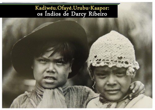 Título da foto: Kadiwéu.Ofayé.Urubu-Kaapor, Índios de Darcy Ribeiro. Foto com duas crianças índias, não de corpo inteiro, apenas do tronco para cima. A da esquerda veste um de chapéu escuro e perpassa o braço em volta da criança da direita, a qual veste uma touca clara. Nota-se um lenço em volta do pescoço de ambas. Cada uma olha para uma direção diferente.