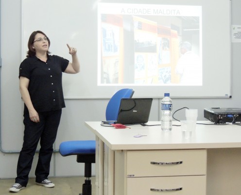 A servidora Rita apresentando um projeto desenvolvido na Justiça Federal Gaúcha. Atrás dela uma imagem projetada no quadro com o título "A Cidade Maldita". ,mais à frente para o lado direito, uma mesa com gavetas e em cima um notebook, projetor e garrafinha de água e copos plásticos.