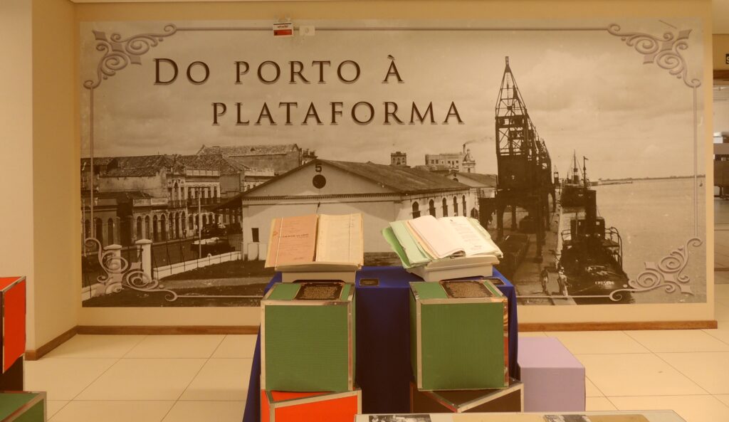 Uma parede com uma imagem grande ,em preto e branco, de um Porto. Título da imagem: "Do Porto à Plataforma". À frente, 4 cubos coloridos (verde, vermelho, marrom) sobrepostos. Atrás desses cubos uma mesa com manto azul ; sobre esta, dois processos abertos em um expositor.