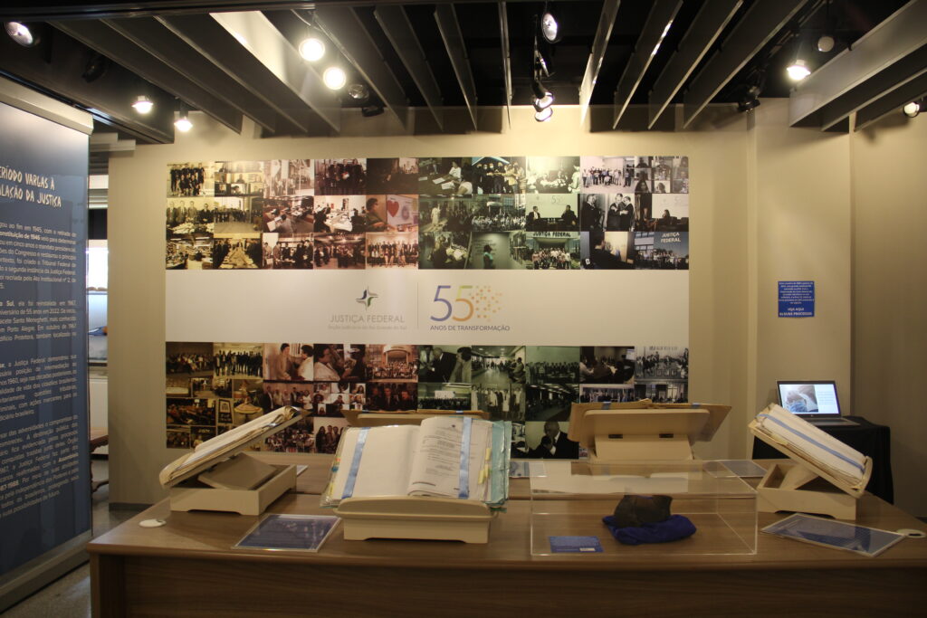 Destaque para mosaico de fotos de servidores e magistrados retratando momentos da história dos 55 anos de reinstalação da JFRS, mesa com processos históricos em exposição