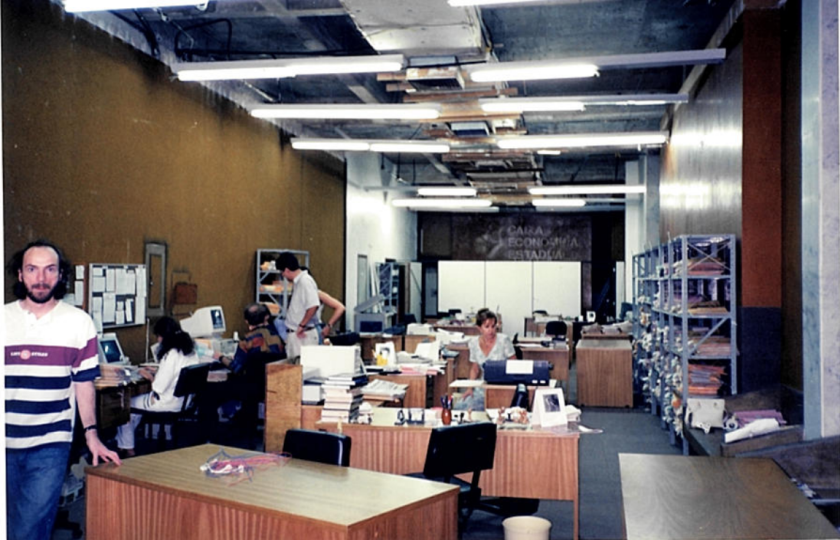a foto antiga mostra uma sala com mesas de trabalho de madeira, pessoas trabalhando nelas, estantes com processos físicos